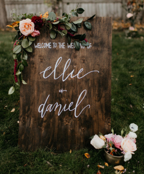 Placa de caligrafia de casamento em madeira que diz Bem-vindo ao casamento de Ellie e Daniel com arranjo de flores