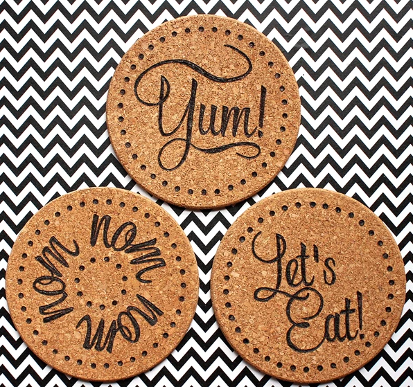 Three wood burned cork trivets each cork trivets has a text on it. 1st says Yum! 2nd says nom nom nom and the 3rd one says Let's eat!